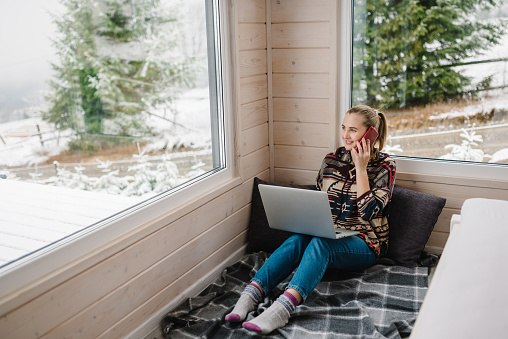 Woman talking on phone. Female remote work on laptop in front of panoramic window with great view of mountain. Remote work from cozy place. Workplace in house in snowy morning. Wide interior view.