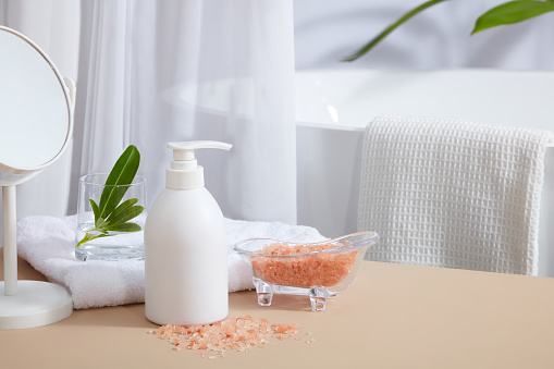 Concept of bathroom with a empty label white bottle decorated with a glass bathtub containing pink himalayan salt and some supplies. Mockup of skin care cosmetic bottle of beauty facial