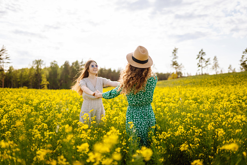 Cheerful girlfriends are walking in a blooming rapeseed field in sunny weather. Two young women enjoying and having fun outdoors. Nature and fun concept.