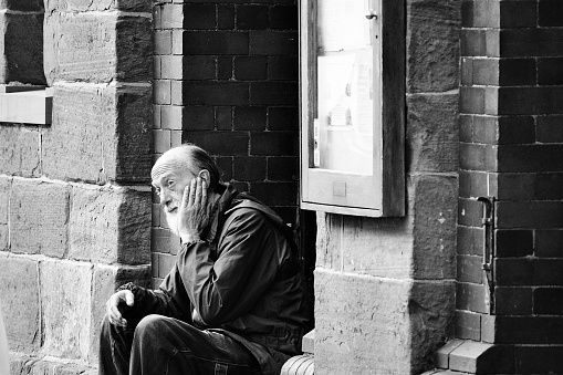 Brecon, Powys, Wales Oct 22 2023: Grey haired old homeless man with a full beard sitting in the doorway of the Guild Hall Council Offices. He is in shabby clothes and looks lost, vacant and confused.