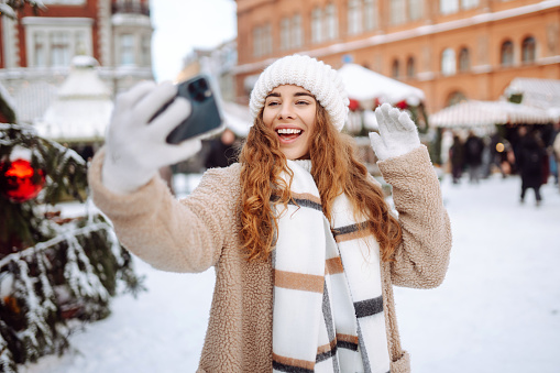 Beautiful young woman having fun and taking a selfie at the New Year's fair. She is dressed in a fur coat, hat, and scarf. Great New Year's mood. The concept of holidays, weekends, relaxation.