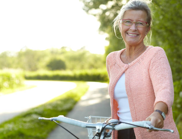 She enjoys healthy outdoor activities - Cycling Smiling senior woman standing on a country lane with her bicycle active seniors summer stock pictures, royalty-free photos & images