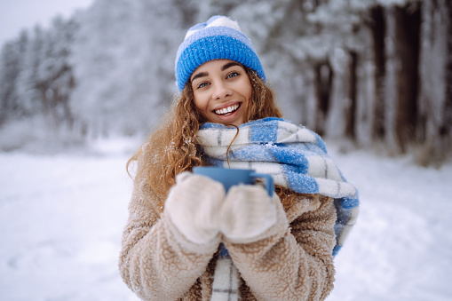 A young woman with a thermos and a cup of coffee in her hands enjoys the first snow in the winter forest. Vacation concept, nature.