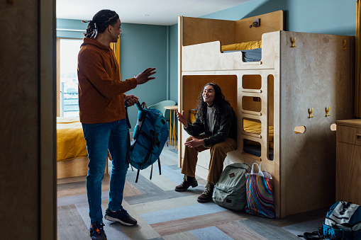 A wide shot of a man and woman wearing warm, casual clothing in a luxury hostel in the seaside town of Amble, Northumberland. They have checked into a dormitory room with bunkbeds. The woman sits on her bed as the man walks towards the door with his backpack in hand and they wave at each other.