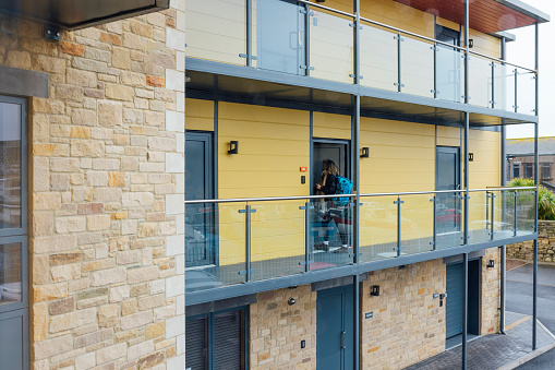 A wide shot of a woman wearing warm, casual clothing on the balcony of a luxury hostel in the seaside town of Amble, Northumberland. She is carrying her backpack and arriving at her room door.