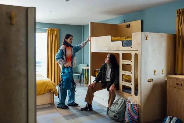 A wide shot of two women wearing warm, casual clothing in a luxury hostel in the seaside town of Amble, Northumberland. They have checked into a dormitory room with bunkbeds. One of them sits on the bed and the other stands, holding her backpack as they talk.