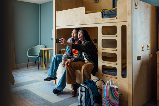 A wide shot of two women wearing warm, casual clothing in a luxury hostel in the seaside town of Amble, Northumberland. They have checked into a dormitory room with bunkbeds. They sit together on one of the beds and take a selfie on a smartphone.