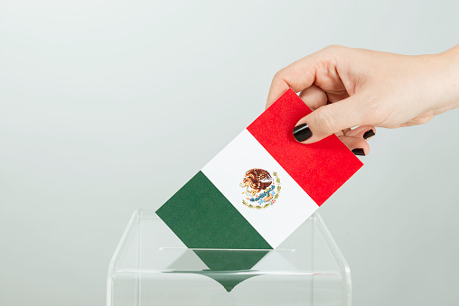 Human hand with black nail polish is inserting flag of Mexico into ballot box.