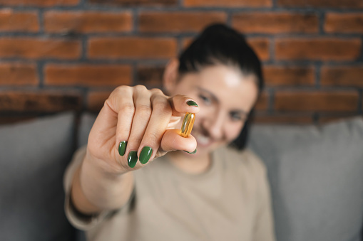 Happy smiling attractive woman recommended omega-3 fish oil supplement, vitamin yellow capsule in her hand, sitting on the sofa at home.