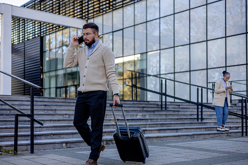 A man, suitcase ready, strides forward with intent, as his black female coworker, stationed a pace behind, taps on her phone, each enacting the familiar rhythm of a workday's commencement.