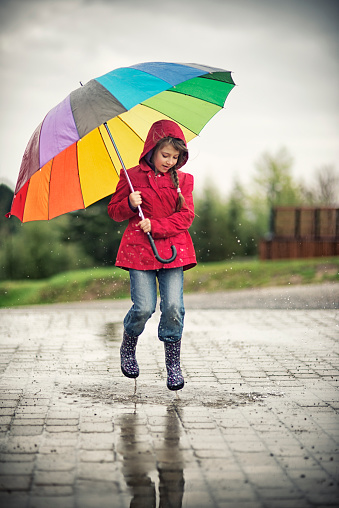 Little girl with colorful umbrella having fun jumping in a puddle. Slightly soft.
