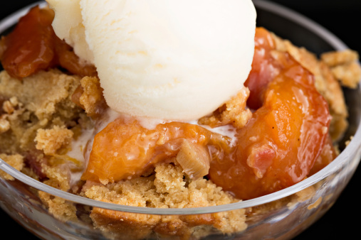 A high angle close up shot of a glass bowl with some hot peach and rhubarb cobbler and a scoop of vanilla ice cream.