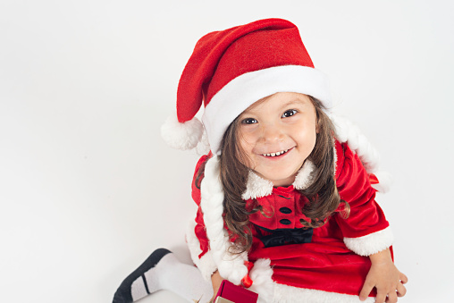 Little girl in Santa Claus costume sitting on white background is looking up at camera with a toothy smile.
