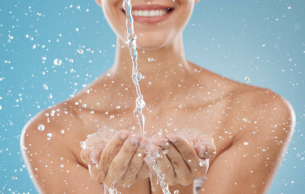 Skincare, water and hands on blue background in studio for hydration, refreshing and facial cleanse. Splash, wellness and woman ready to clean face for washing, moisturizing and hydrate healthy skin