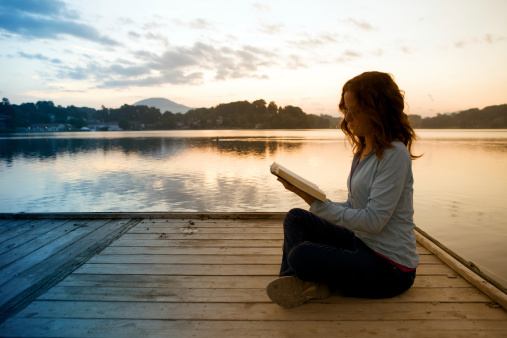 Young woman reading the Bible by a lake at sunrise
