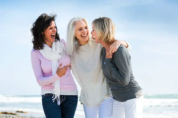 Portrait of three mature women having fun together at beach. Women are laughing out loud with arms are around each other. They have been best friends for years. The blue sky and ocean in the background. Women are between 50 and 65 years old. Girlfriends feel happy and confident at their stage of life. Nobody can make you laugh like your best friends. 