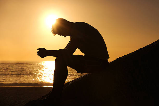 Burdened by unseen worries Silhouette of a man sitting by the ocean and contemplating while looking downwards one man only stock pictures, royalty-free photos & images