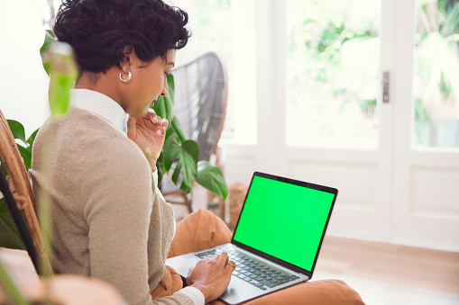 A young woman is captured in her living room, diligently working on her laptop with a green screen, her focused posture a testament to her dedication. This glimpse into her workspace reveals a blend of professionalism and the coziness of home, highlighting the evolving nature of modern remote work