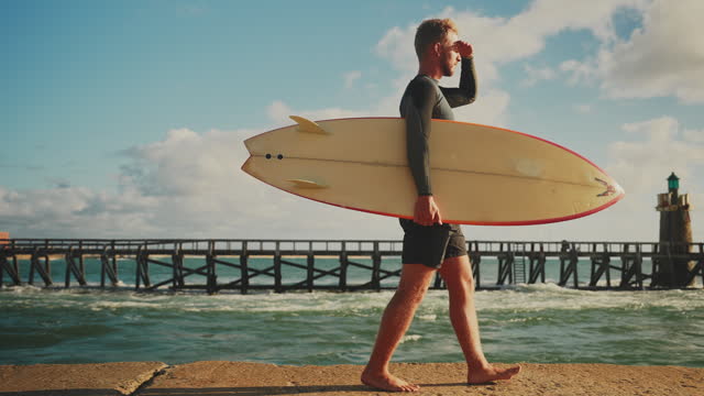 Male surfer walking with surfboard along sunny ocean pier