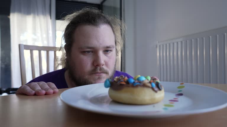 One overweight young man sitting in front of donut refraining from eating sugar sweet unhealthy snack. Temptation concept