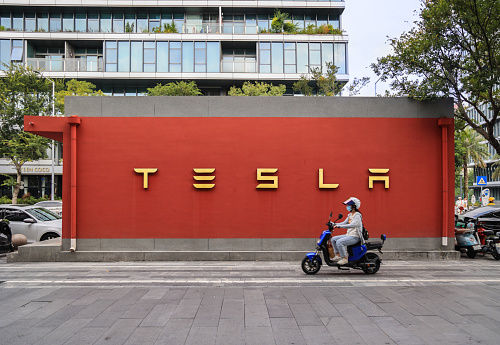 Shenzhen, China - November 06.2023: The new energy vehicle Tesla billboard on a red wall with a passing motorbike in front, Shenzhen China