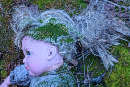 part of an old dirty plastic toy doll with a head with gray hair overgrown with green moss lies on the ground on the street