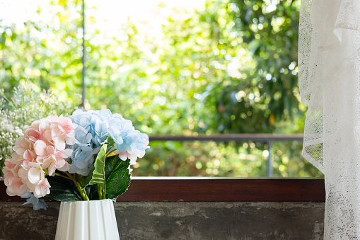 Beautiful and cozy style background with copy space of colorful flowers in vase with wooden frame of window in vintage or retro decoration seeing natural summer sunlight on greenery trees and leafs.