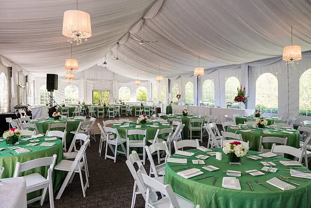 The inside of a fancy outdoor party tent prepared with sheer fabric draped ceilings, round guest tables, folding chairs, table cloths, centerpieces and flowers for a birthday party.