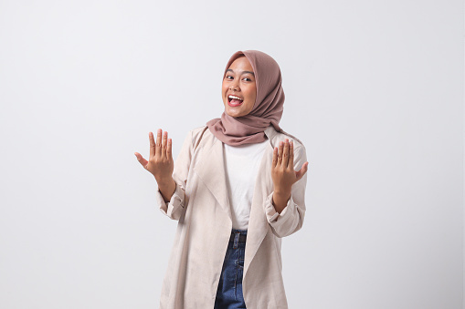 Portrait of excited Asian hijab woman in casual suit spreading her hands sideways. Greeting and welcoming someone. Inviting people to come in. Businesswoman concept. Isolated image on white background