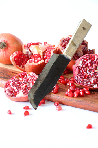 Stock photo showing a close-up view of healthy eating image of a wooden chopping board covered in pomegranates (Punica granatum) in various states of being cut up. Whole, halved and quartered pieces of fruit displaying red-pink peel (epicarp), white mesocarp (albedo) and red flesh (arils) encasing seeds against a white background.