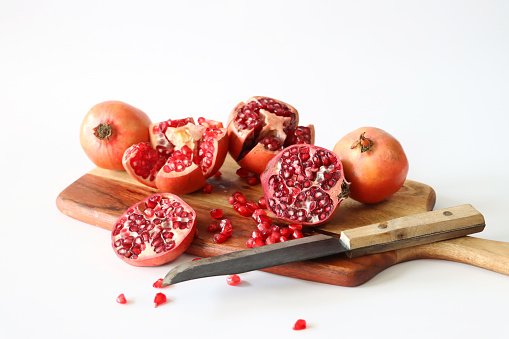 Stock photo showing a close-up view of healthy eating image of a wooden chopping board covered in pomegranates (Punica granatum) in various states of being cut up. Whole, halved and quartered pieces of fruit displaying red-pink peel (epicarp), white mesocarp (albedo) and red flesh (arils) encasing seeds against a white background.