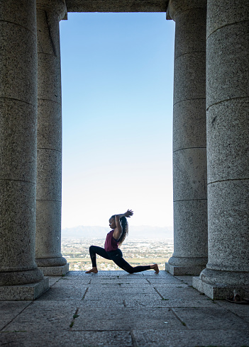 Fit young woman in sportswear practicing the crescent lunge pose by stone columns of a monument during an outdoor yoga session