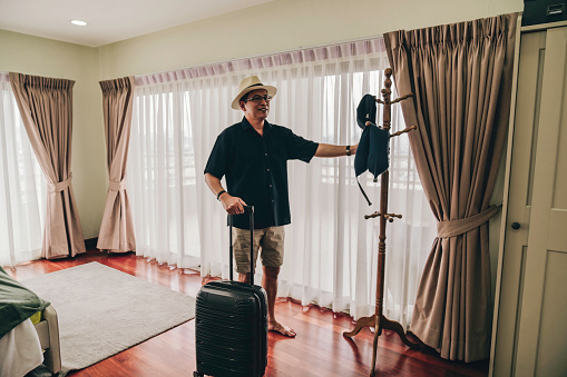 senior man standing with luggage by bed in hotel room during weekend