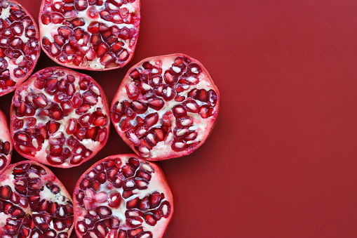 Stock photo showing a close-up, elevated view of healthy eating image of pomegranates (Punica granatum) halves. Pieces of fruit displaying red-pink peel (epicarp), white mesocarp (albedo) and red flesh (arils) encasing seeds on a red background.