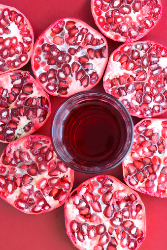 Stock photo showing close-up, elevated view of healthy eating image of a drinking glass of red pomegranate juice besides slices of pomegranate fruit displaying red-pink peel (epicarp), white mesocarp (albedo) and red flesh (arils) encasing seeds on a red background.