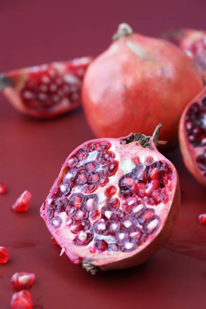 Stock photo showing a close-up, elevated view of healthy eating image of pomegranates (Punica granatum) in various states of being cut up. Whole, halved and quartered pieces of fruit displaying red-pink peel (epicarp), white mesocarp (albedo) and red flesh (arils) encasing seeds. imagen de granadas (punica granatum), rodajas de fruta enteras, cortadas por la mitad y en cuartos que muestran cáscara de color rojo rosado (epicarpio), mesocarpio blanco (albedo) y pulpa roja (arilos) que envuelven semillas, fondo rojo, enfoque en prime