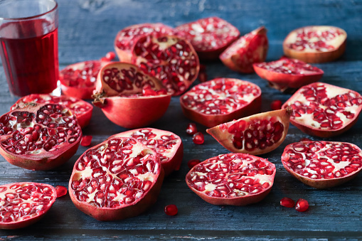 Stock photo showing close-up, elevated view of healthy eating image of a drinking glass of red pomegranate juice besides halves, quarters and slices of pomegranate (Punica granatum) fruit displaying red-pink peel (epicarp), white mesocarp (albedo) and red flesh (arils) encasing seeds on a blue wood grain background.