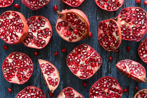 Stock photo showing a close-up, elevated view of healthy eating image of pomegranates (Punica granatum) whole, halved, quartered and sliced fruits. Pieces of fruit displaying red-pink peel (epicarp), white mesocarp (albedo) and red flesh (arils) encasing seeds on a blue wood grain background.