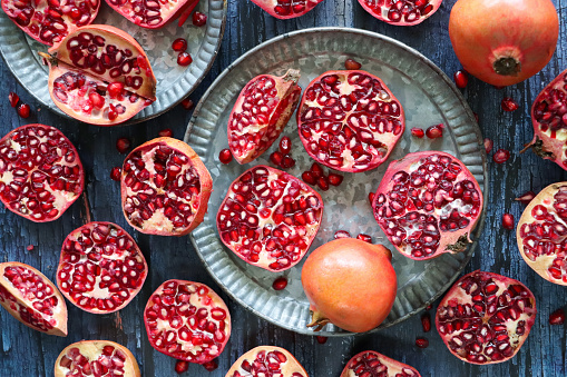 Stock photo showing a close-up, elevated view of healthy eating image of pomegranates (Punica granatum) whole, halved, quartered and sliced fruits on a grey plate. Pieces of fruit displaying red-pink peel (epicarp), white mesocarp (albedo) and red flesh (arils) encasing seeds on a blue wood grain background.