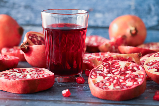 Stock photo showing close-up view of healthy eating image of a drinking glass of red pomegranate juice besides halves, quarters and slices of pomegranate (Punica granatum) fruit displaying red-pink peel (epicarp), white mesocarp (albedo) and red flesh (arils) encasing seeds on a blue wood grain background.