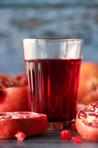 Stock photo showing close-up view of healthy eating image of a drinking glass of red pomegranate juice besides halves, quarters and slices of pomegranate (Punica granatum) fruit displaying red-pink peel (epicarp), white mesocarp (albedo) and red flesh (arils) encasing seeds on a blue wood grain background.