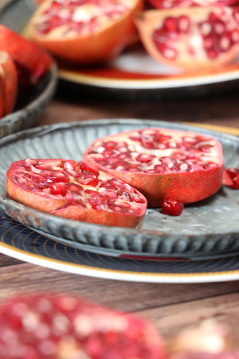 Stock photo showing a close-up view of healthy eating image of pomegranates (Punica granatum) whole, halved, quartered and sliced fruits on a grey plate. Pieces of fruit displaying red-pink peel (epicarp), white mesocarp (albedo) and red flesh (arils) encasing seeds on a wood grain background.