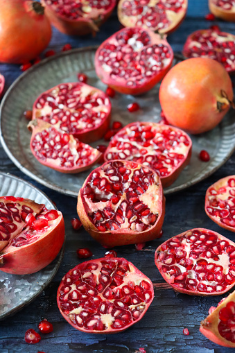 Stock photo showing a close-up view of healthy eating image of pomegranates (Punica granatum) whole, halved, quartered and sliced fruits on a grey plate. Pieces of fruit displaying red-pink peel (epicarp), white mesocarp (albedo) and red flesh (arils) encasing seeds on a blue wood grain background.