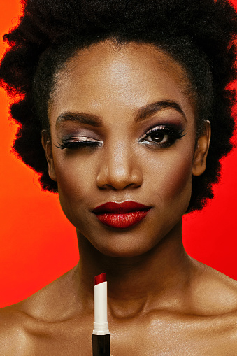 Wink, cute African American woman with curly hair beauty studio shot close up eye holding a red lipstick. Glamour cosmetics advertisement concept