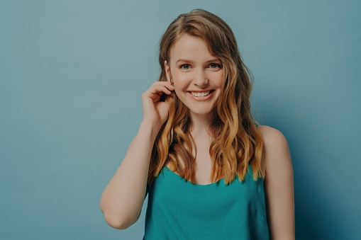 Cheerful young woman with wavy hair smiling, touching ear against a soft blue background