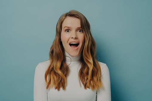 Amazed young woman in a white turtleneck, her mouth open in excitement, against a pale blue background