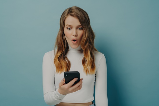 Shocked young woman holding phone, mouth agape, expressing surprise, dressed in white on a teal background