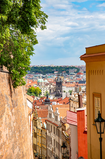 Prague, Czech Republic from a hill. The city is visible in the background, with its red-roofed buildings and a few taller buildings standing out against the blue sky with a few clouds. In the foreground, there is a tree with green leaves, a yellow building with a red roof, and a black street lamp on the right side of the image.