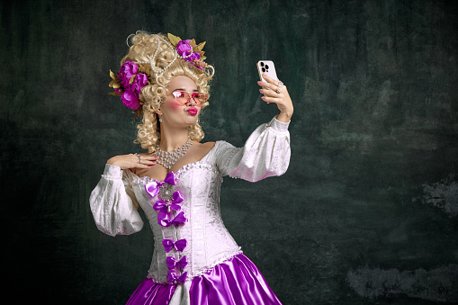 Woman dressed like classic historical character, in old-fashioned dress and taking selfie on mobile phone against vintage background. Concept of technology, blogging, shopping, sales, delivery, online