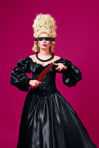 Retrato de una persona medieval vestida con un vestido negro anticuado con una venda calada jugando con un cuchillo de color sobre un fondo magenta. photo